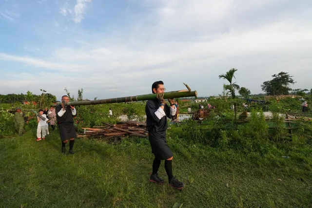 Two men in traditional dress carrying a long bamboo pole across a green field at the Gelephu Mindfulness City site