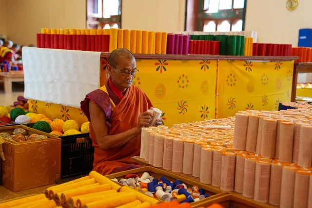 A monk in maroon robes seated among stacks of gold and orange materials in a workshop, preparing offerings