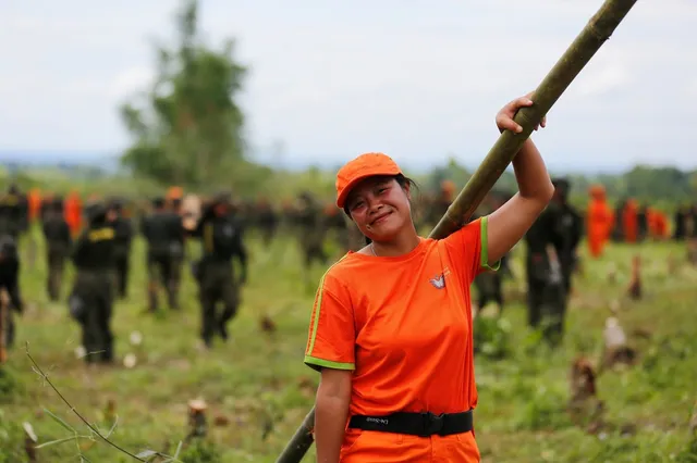 A smiling volunteer in an orange uniform holding a bamboo pole over their shoulder, with others at work on cleared ground behind