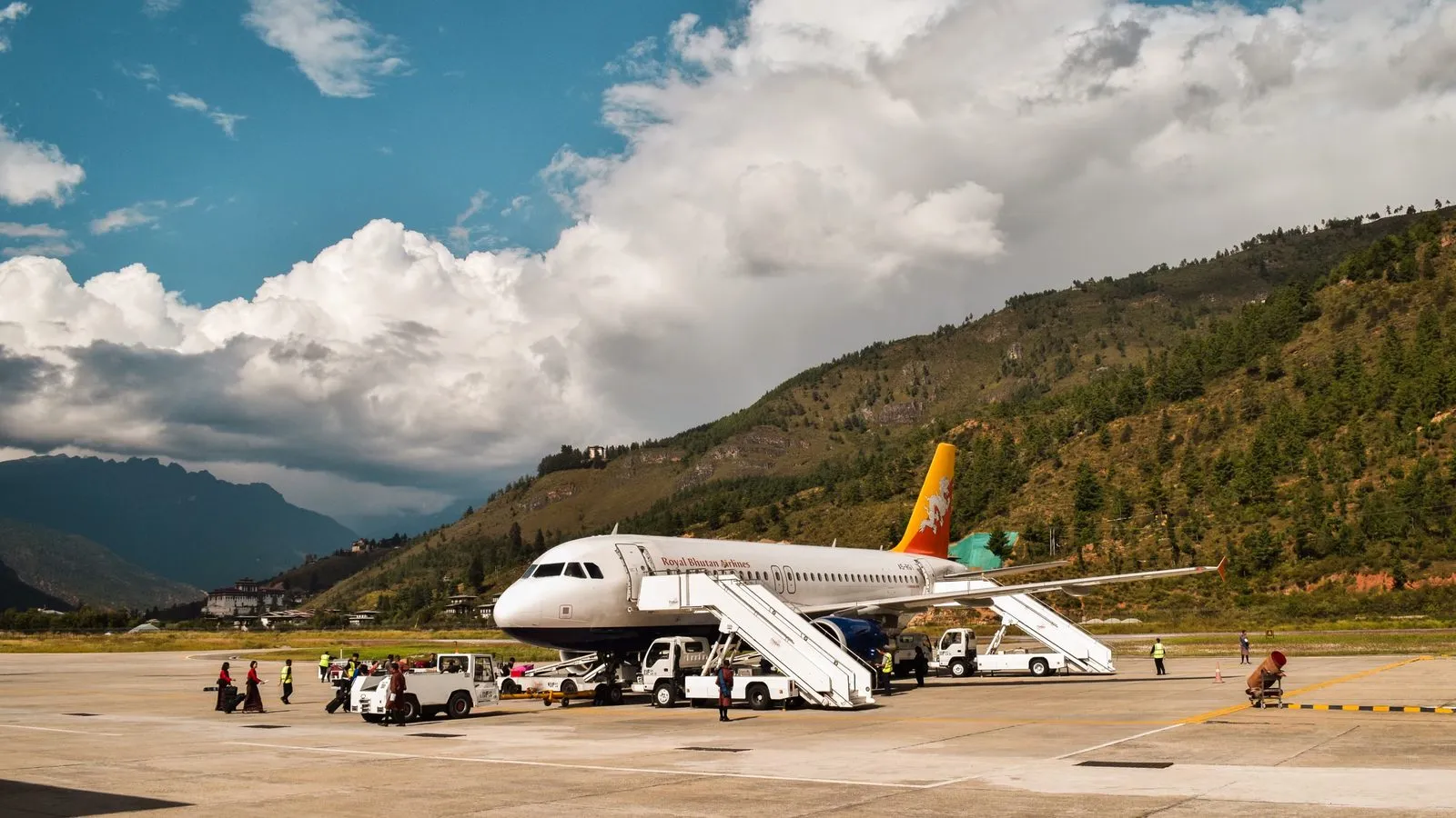 A passenger jet on the apron of a valley airport, with steps against the fuselage and forested hills behind