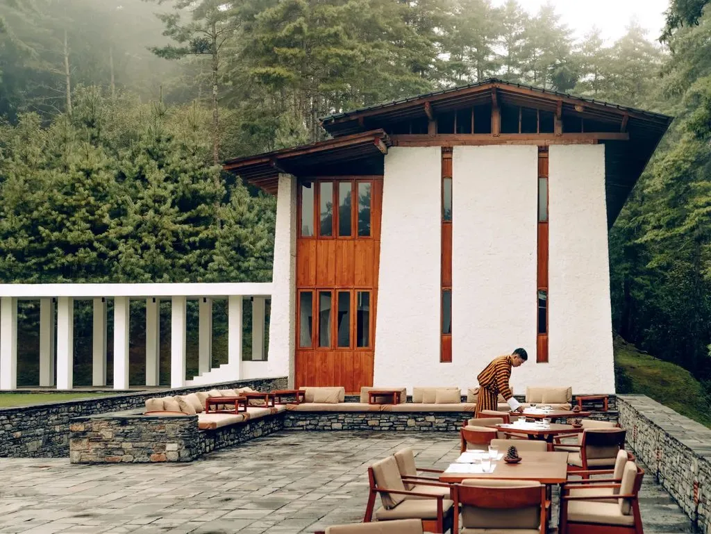 A whitewashed lodge with timber doors and a pitched roof among misty pines, with a terrace set for a meal