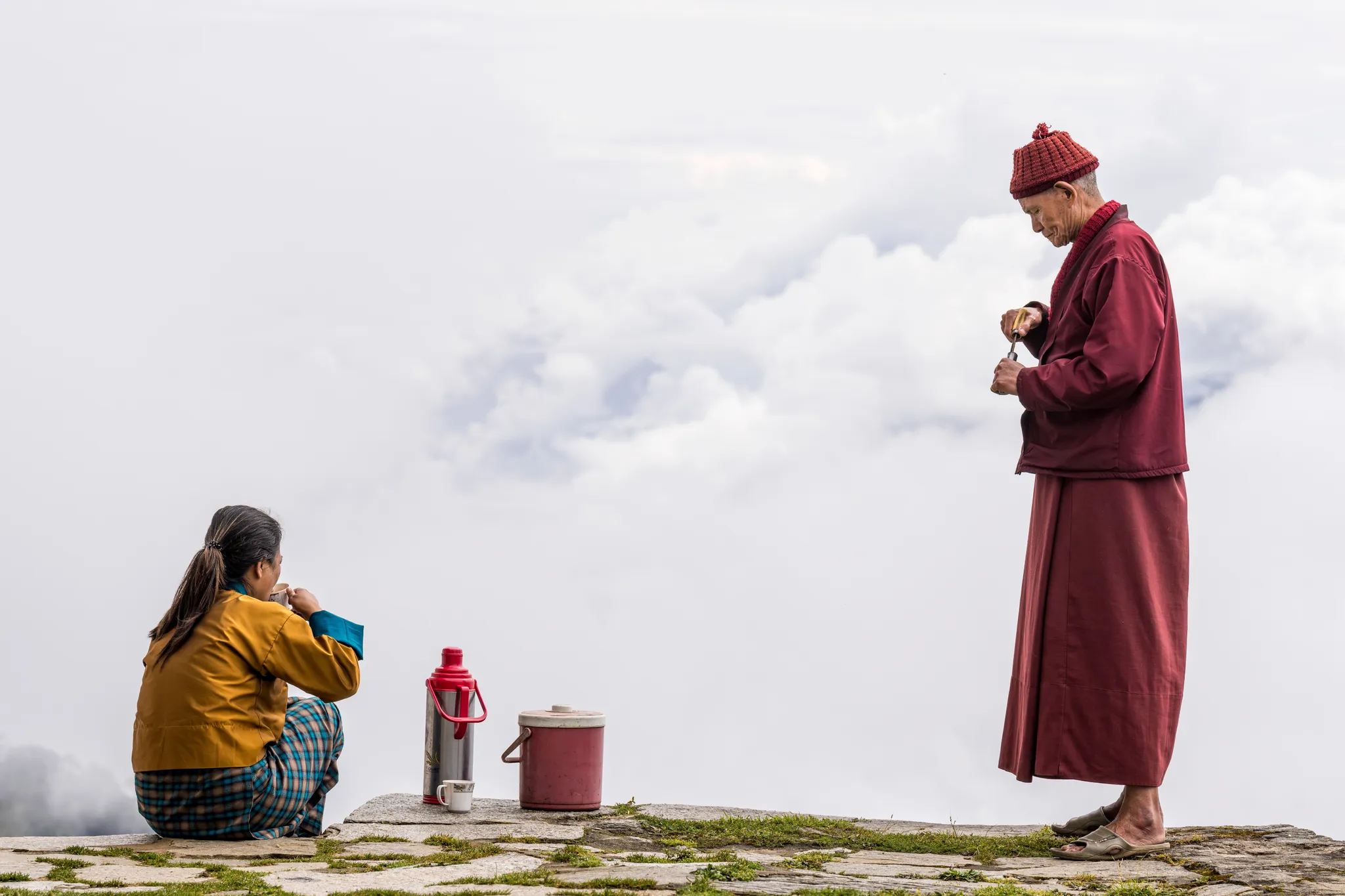 A monk in maroon robes pours from a flask on a stone terrace above the clouds while a woman sits nearby with a cup of tea