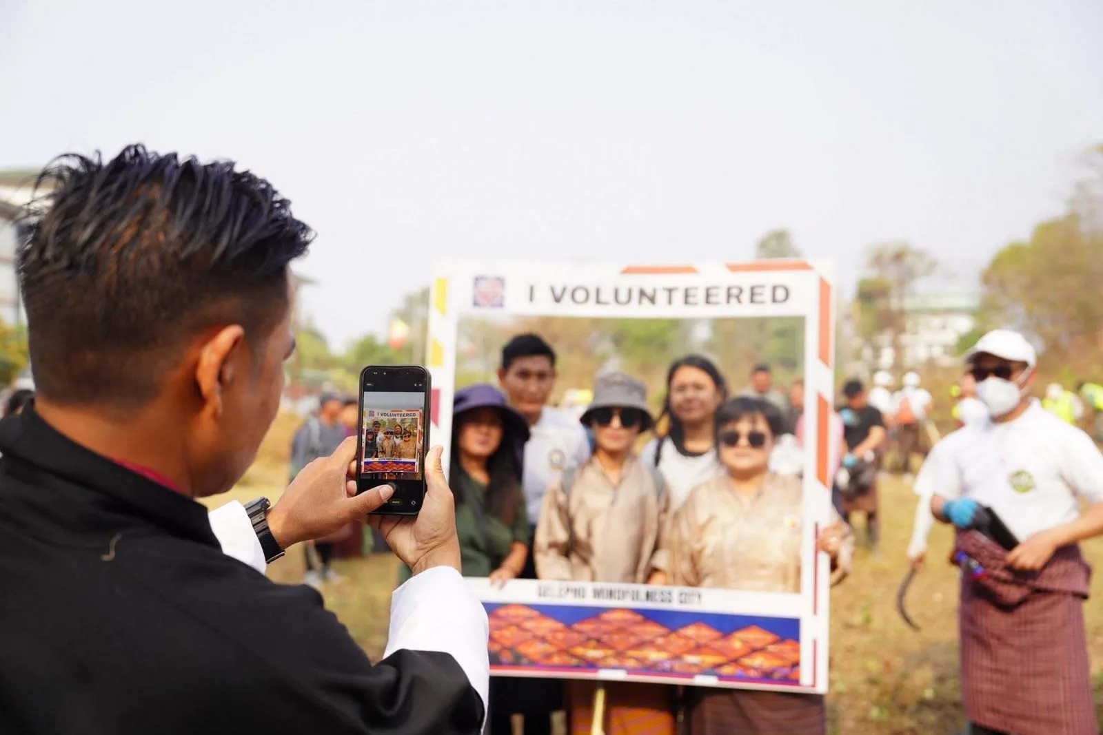 A man photographs a group of volunteers posing behind an "I volunteered" frame in an open field, with other volunteers at work behind them
