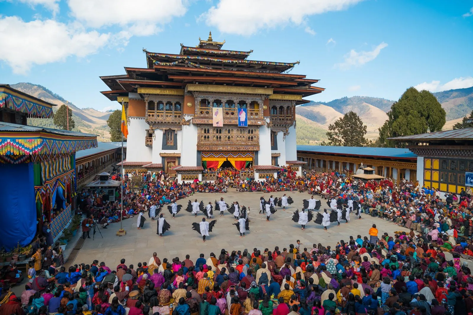 Dancers in black and white winged costumes performing in a monastery courtyard, ringed by a large seated crowd